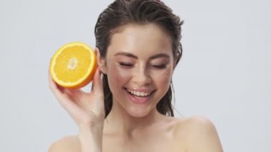 Mutlu, gülümseyen, yarı çıplak, ıslak saçlı bir kadın stüdyoda açık mavi bir arkaplanda portakal tutarken poz veriyor.