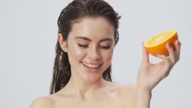 Çekici, yarı çıplak, ıslak saçlı bir kadın stüdyoda açık mavi bir arka planda izole edilmiş portakal meyvesiyle poz veriyor.