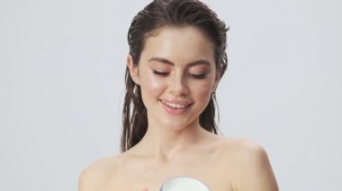 Güzel, yarı çıplak, ıslak saçlı bir kadın omzuna bir vücut kremi sürüyor. Stüdyoda açık mavi bir arka planda izole edilmiş.