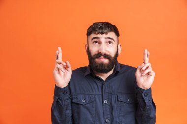 Burun mücevherleri takan, siyah tişörtlü, yukarı bakan, turuncu arka planda parmakları çaprazlanmış genç bir adam resmi.