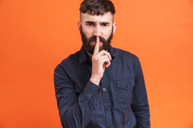 Burnu mücevherlerle dolu genç bir adamın görüntüsü. Parmağını dudaklarına dayayıp turuncu arka planda sessizliğini korumak istiyor.