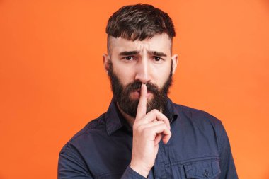 Burnu mücevherlerle dolu genç bir adamın görüntüsü. Parmağını dudaklarına dayayıp turuncu arka planda sessizliğini korumak istiyor.