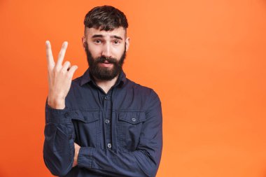 Siyah gömlekli esmer adamın, turuncu arka planda iki parmağını kameraya gösterirken çekilmiş bir fotoğrafı.