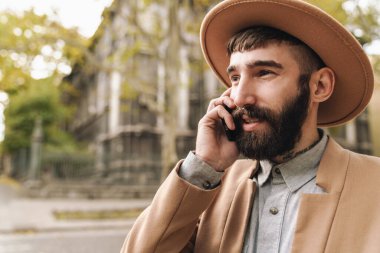 Açık havada yürürken cep telefonuyla konuşan şapkalı çekici bir genç resmi.