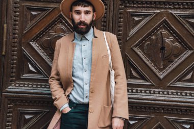 Sonbahar ceketi ve şapkası giyen çekici genç adam şehrin dışında, ahşap kapının önünde dikiliyor.