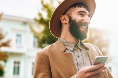 Sonbahar ceketi ve şapkası giyen çekici bir genç adam elinde cep telefonuyla şehir caddesinde yürüyor.