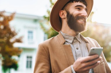Sonbahar ceketi ve şapkası giyen çekici bir genç adam elinde cep telefonuyla şehir caddesinde yürüyor.