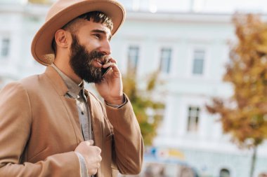 Sonbahar ceketi ve şapkası giyen çekici genç adam şehir caddesinde yürüyor, cep telefonuyla konuşuyor.