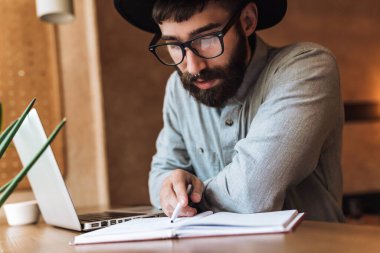 Gözlük takan, dizüstü bilgisayar kullanan ve kafede otururken kitap okuyan genç bir adamın fotoğrafı.