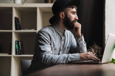 Ciddi bir genç adamın kafeteryada otururken dizüstü bilgisayarında daktilo kullanmasının fotoğrafı.