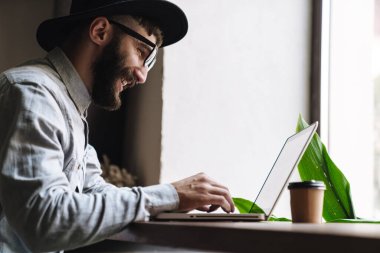 Gülen gözlüklü genç adamın bilgisayarda yazı yazarken ve kafede otururken kahve içerken çekilmiş fotoğrafı.