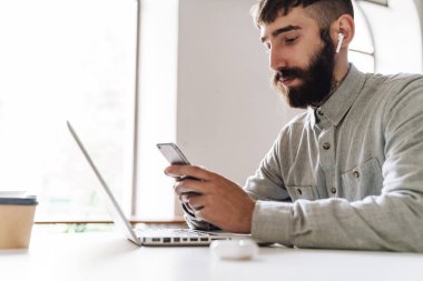 Ofiste otururken cep telefonu ve kulaklık kullanan piercing takan ciddi bir gencin fotoğrafı.