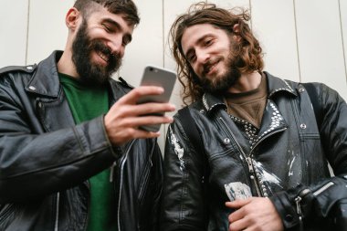 Deri ceketli, sakallı, neşeli adamların fotoğrafı. Cep telefonu kullanıyor ve dışarıda duvara yaslanırken gülümsüyor.