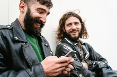 Deri ceketli, sakallı, neşeli adamların fotoğrafı. Cep telefonu kullanıyor ve dışarıda duvara yaslanırken gülümsüyor.