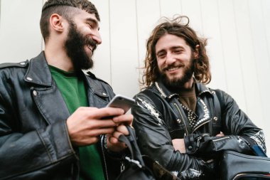 Deri ceketli, sakallı, neşeli adamların fotoğrafı. Cep telefonu kullanıyor ve dışarıda duvara yaslanırken gülümsüyor.