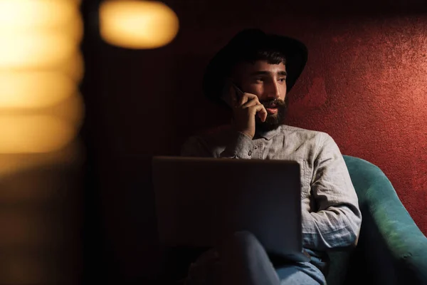 Foto Jovem Barbudo Usando Chapéu Usando Laptop Falando Celular Enquanto — Fotografia de Stock