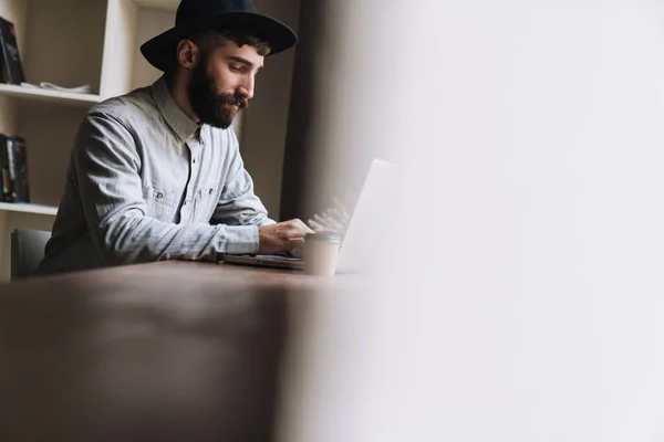 Foto Jovem Sério Usando Chapéu Digitando Laptop Bebendo Café Enquanto — Fotografia de Stock