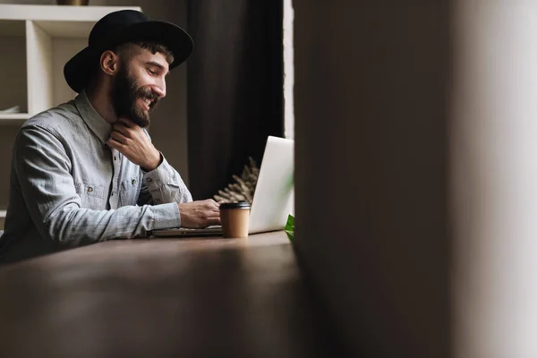 Photo Laughing Young Man Wearing Hat Typing Laptop Drinking Coffee — Stock Photo, Image