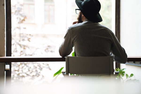 Фото молодого человека в очках, смотрящего в окно, сидящего за столом в кафе в помещении
