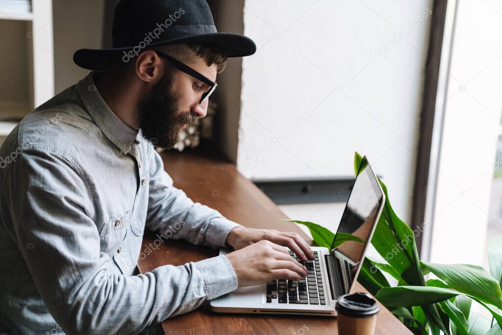 Photo of thinking young man wearing eyeglasses typing on laptop and drinking coffee while sitting at table in cafe indoors