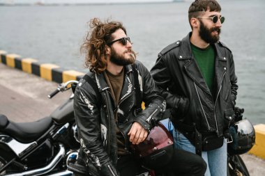 Bisikletleriyle deniz limanında dikilirken konuşan beyaz sakallı motorcuların fotoğrafı.