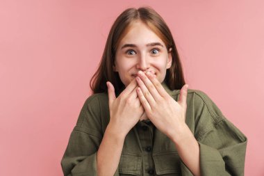 Heyecanlı beyaz kadının ağzını kapattığı ve pembe arka planda izole edilmiş kameraya baktığı fotoğraf.