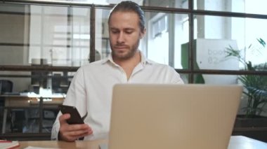 Ofisteki genç ve yakışıklı iş adamı dizüstü bilgisayar ve cep telefonu kullanıyor.