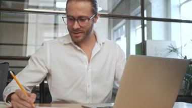 Ofisteki yakışıklı iş adamı dizüstü bilgisayar kullanarak not tutuyor.