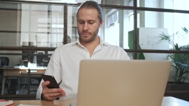 在办公室里用手提电脑和手机集中精神的年轻英俊商人 — 图库视频影像