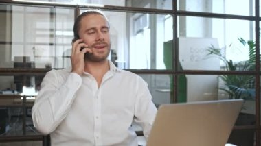 Ofiste cep telefonuyla konuşan dizüstü bilgisayar kullanan memnun genç bir iş adamı.