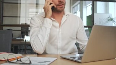 Ofisteki ciddi genç iş adamı cep telefonuyla konuşan dizüstü bilgisayarını kullanıyor.