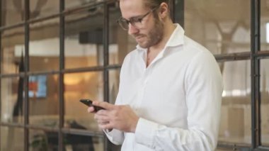 Yakışıklı memnun genç iş adamı ofiste cep telefonuyla yürüyor.