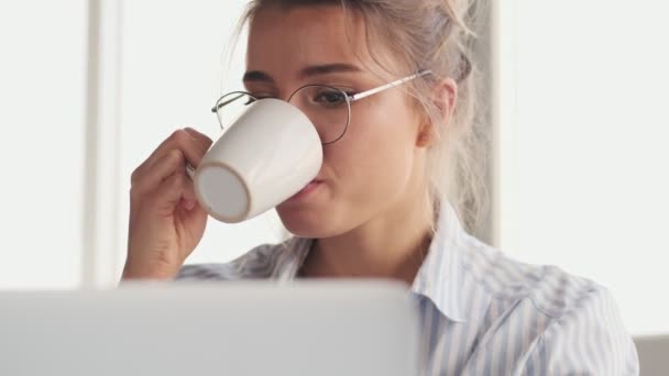 ガラスを身に着けている美しい若い女性は家に座っている間ノートパソコンで作業中にコーヒーを飲んでいます — ストック動画