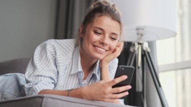 Neşeli genç bir kadın akıllı telefonuyla komik bir şey izlerken evinde kanepede oturuyor.