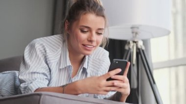 Çekici genç bir kadın akıllı telefonuyla komik bir şey izlerken kanepede oturuyor.
