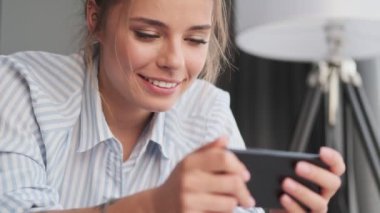 Oldukça hoş bir genç bayanın yakın plan görüntüsü akıllı telefonuyla evinde kanepede otururken onu yatay olarak tutarken bir şey izliyor.