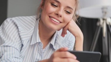 Gülümseyen genç bir kadının yakından görüntüsü, akıllı telefonuyla evdeki kanepede yatay olarak uzanmış bir şekilde bir şey izliyor.