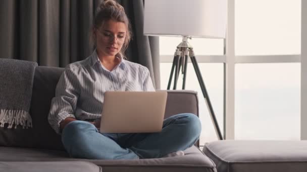 一位戴着眼镜的年轻貌美的女士正坐在家里的沙发上用她的笔记本电脑 — 图库视频影像