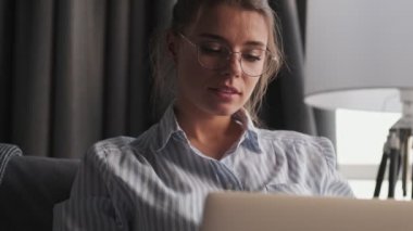 Gözlüklü çekici bir genç kadın dizüstü bilgisayarını kullanıyor ve oturma odasında otururken yan tarafa bakıyor.