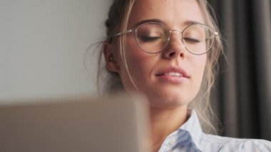 Gözlüklü güzel bir genç kadının yakın plan görüntüsü evinde otururken dizüstü bilgisayarını kullanıyor.