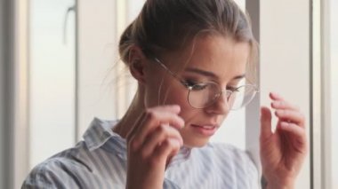 Güzel, sakin genç bir kadın, oturma odasında penceresinden dışarı bakarken gözlüklerini çıkarıyor.