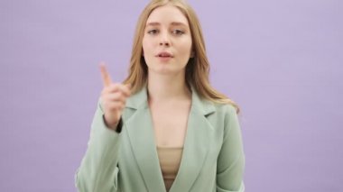 Gülümseyen genç bir kadın, gri arka planda tek başına dururken sessiz bir jest yapıyor.