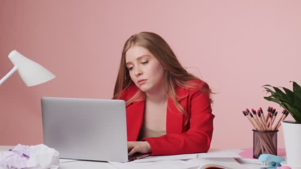 一位非常疲倦的年轻女士正在用她的笔记本电脑工作 她坐在桌子旁 与粉红的背景隔离 — 图库视频影像