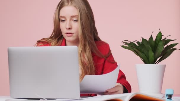 A concentrated young woman is typing on her laptop computer and working with paper documents sitting at the table isolated over pink background