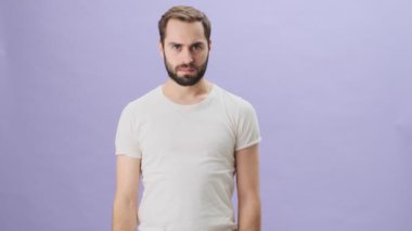 Beyaz t-shirt giyen ciddi bir genç adam gri arka planda tek başına dururken ellerini çapraz yapıyor.