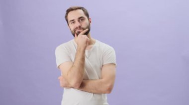 Sakin, beyaz tişört giyen genç bir adam gri arka planda tek başına dururken bir şey düşünüyor.