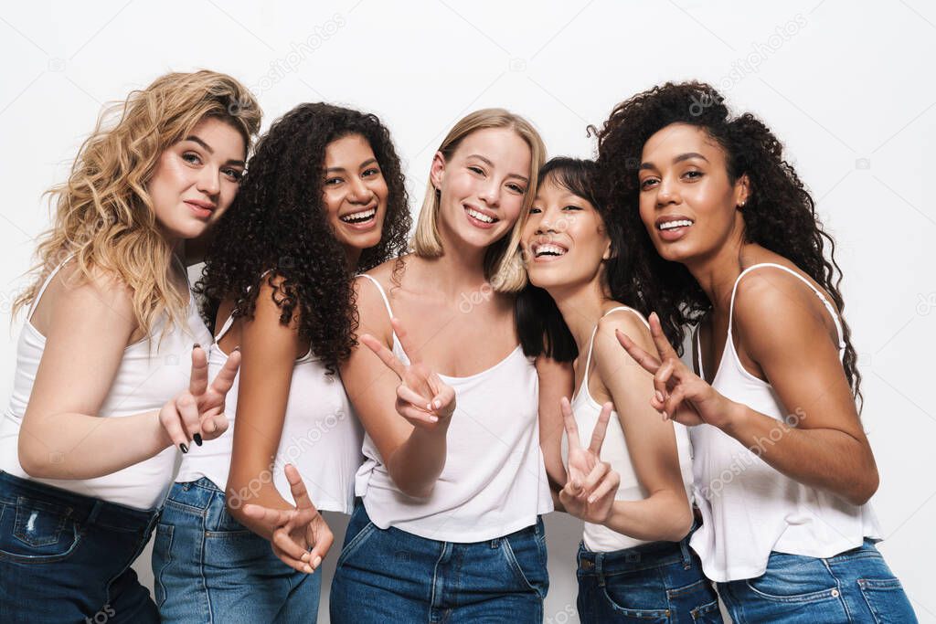 Image of pretty multinational women in blue jeans smiling and gesturing peace sign isolated over white background
