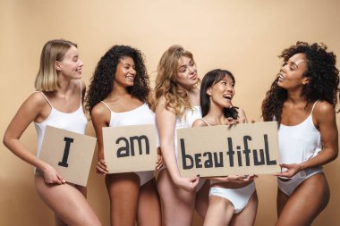 İç çamaşırlı, gülümseyen ve bej arka planda izole edilmiş pankartlar tutan çok uluslu genç kadınların görüntüsü