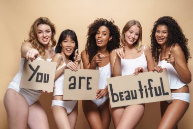 Image of funny multinational women in underclothes pointing fingers at camera and holding placards isolated over beige background clipart