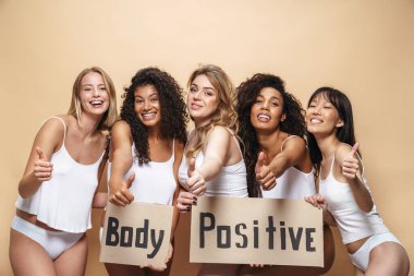 İç çamaşırlı, mutlu, çok uluslu kadınların bej arka plan üzerinde başparmaklarını gösteren ve pankartlarını tutan resimleri.